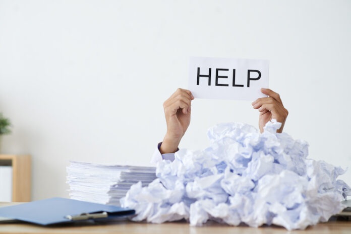 Person under heap of crumpled papers with hand holding a help sign