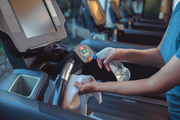 Gym cleaning and disinfection. Infection prevention and control of epidemic. Staff using wipe and alcohol sanitizer spray to clean treadmill in gym. Anti Covid-19 precautions
