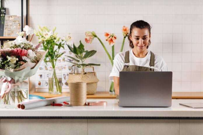 Young flower shop owner using laptop at counter. Woman florist standing at table.